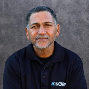 Portrait of a smiling middle-aged man with short gray hair and a light beard, wearing a dark polo shirt with an ICSolar logo against a gray textured backdrop.