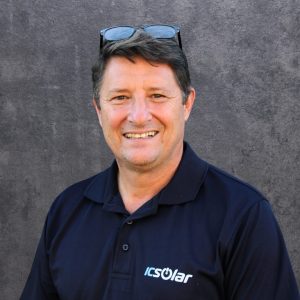 Smiling man in a navy polo with the ICSolar logo, sunglasses resting on his head, standing against a gray textured wall.