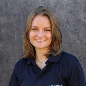 Portrait of a smiling woman with shoulder-length brown hair, wearing a navy polo shirt, against a gray wall.