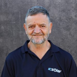 Portrait of a smiling, middle-aged man with short gray hair and beard, wearing a navy polo shirt with an ICSolar logo, against a gray textured backdrop.