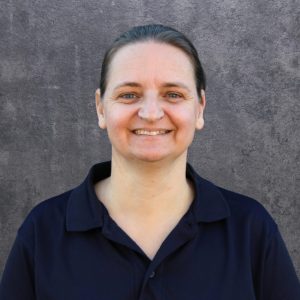 Smiling woman with dark hair tied back, wearing a navy polo shirt, posed against a gray textured wall outdoor or studio backdrop.