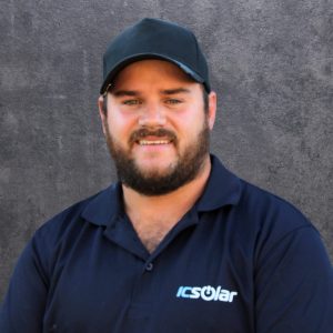 Portrait of a smiling man wearing a navy ICSolar polo shirt and a dark cap, standing against a gray textured wall.